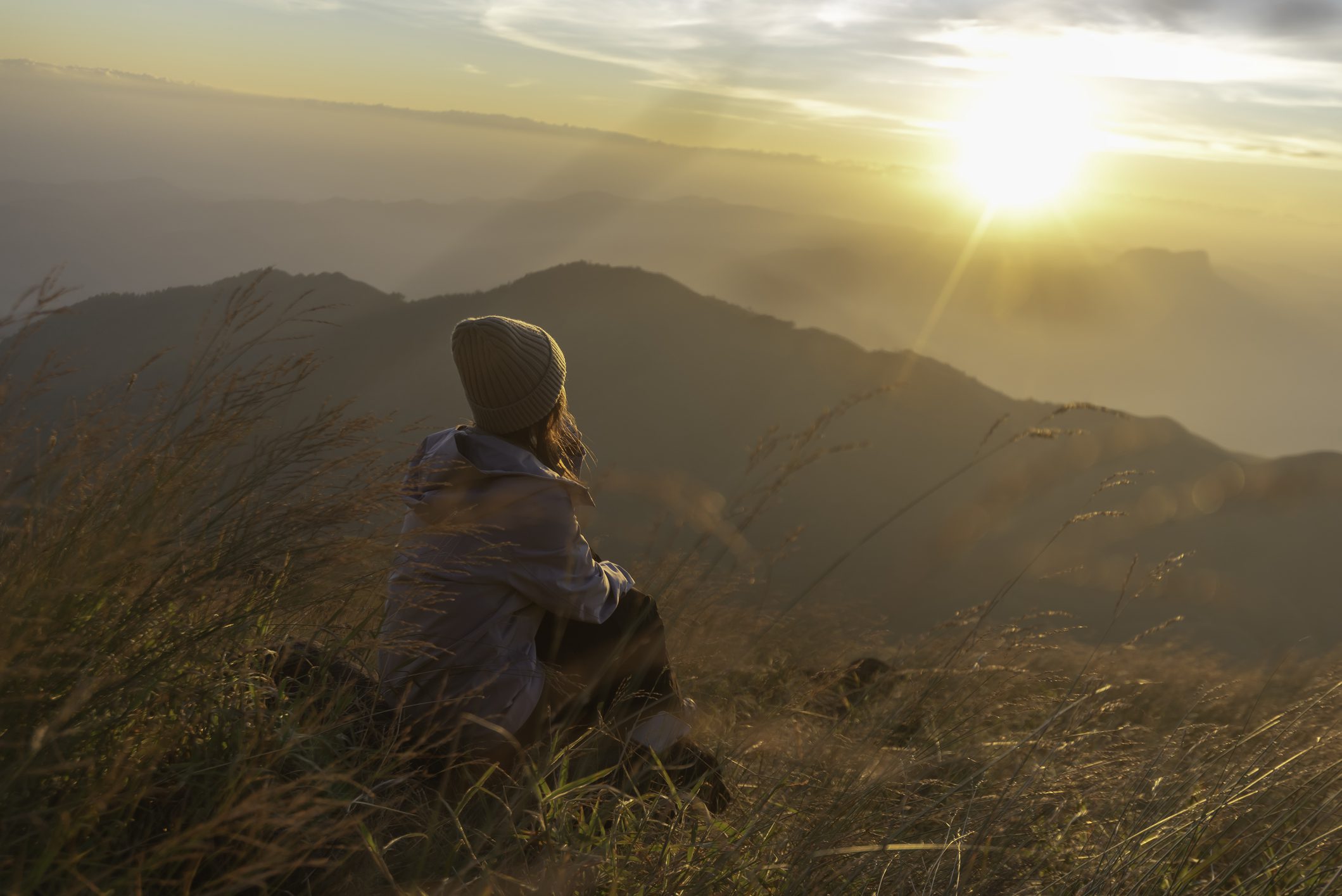 Person sitting on a grassy hill watching the sunrise over mountains.